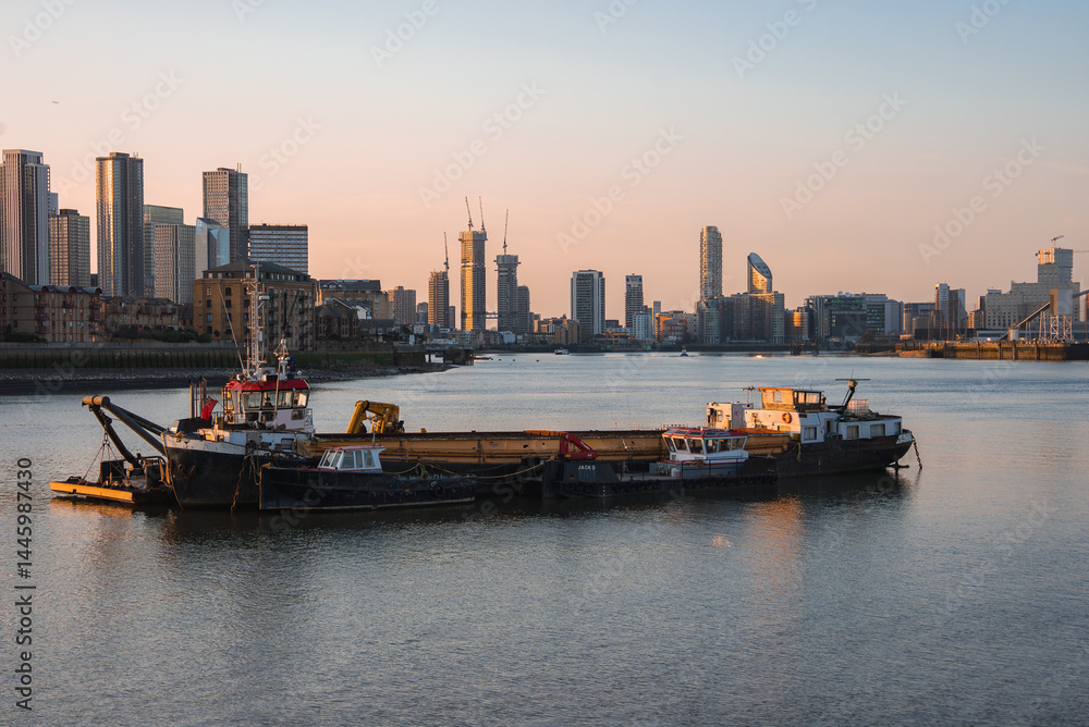 View of the River Thames at sunset in London, featuring anchored boats, a barge, and the modern skyscrapers of Canary Wharf reflecting warm light.