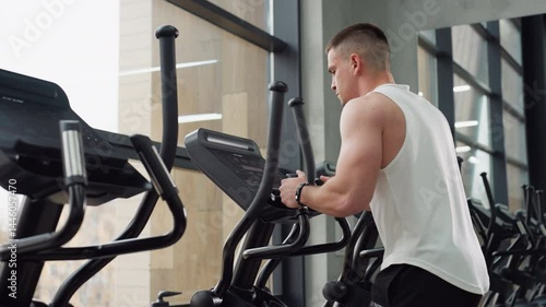Side view of gym junkie on elliptical trainer gripping handles and staring at console, muscles engaged during intense cardio session in bright urban fitness studio with glass windows and equipment