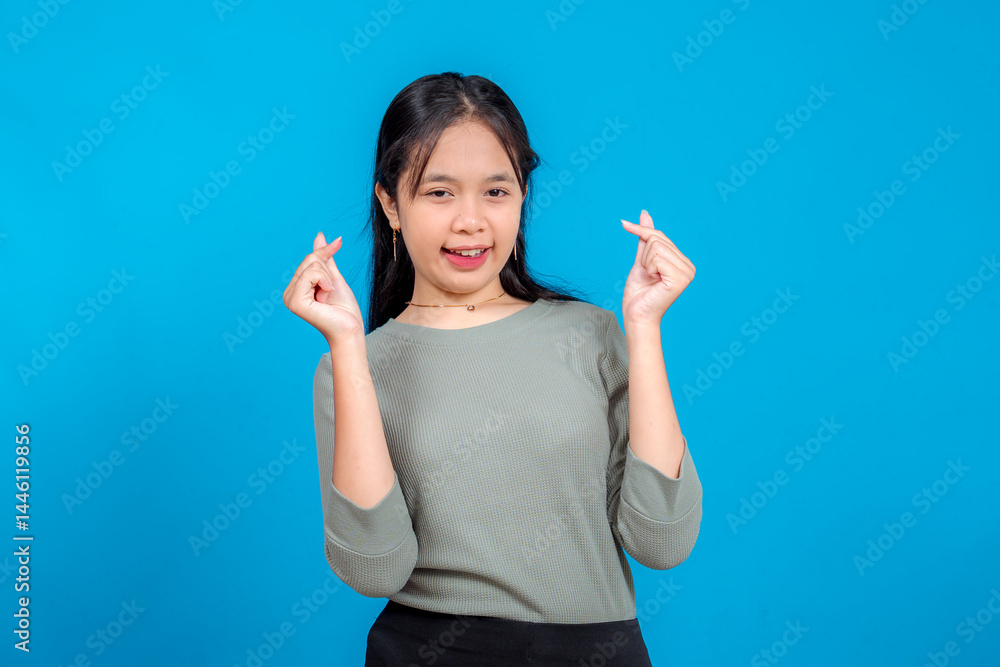 Fototapeta premium Happy young Asian girl smiling and making double finger heart gestures. Her bright expression, casual outfit, and playful charm pop against a clean blue studio background.