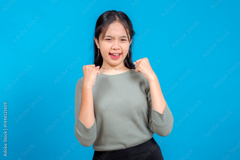Fototapeta premium Excited young Asian woman with a confident and determined facial expression, clenching fists in front of her chest. Isolated on blue background. Concept of success and motivation.
