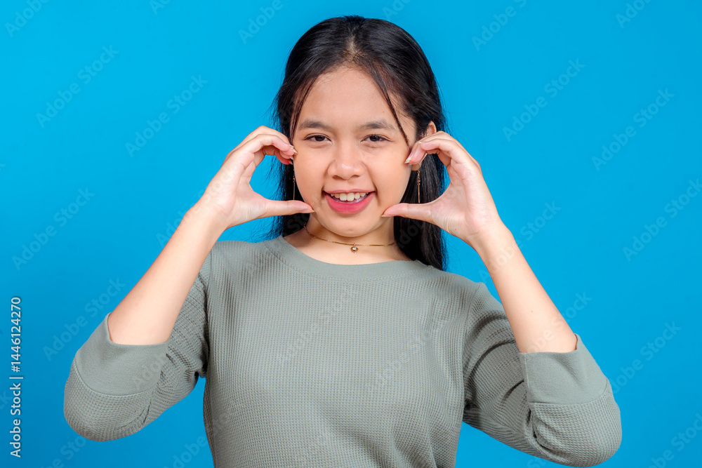 Naklejka premium Cheerful young Asian woman with long hair smiling and playfully framing her cheeks with her hands against a blue background. Joyful facial expression for lifestyle and advertising use.
