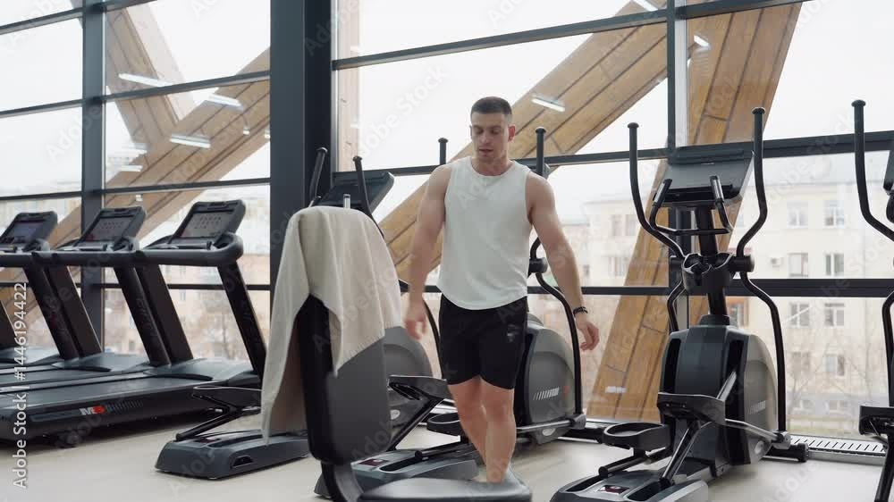 Student steps down from tread mil machine after intense cardio session, looks exhausted while grabbing towel hanging on chair to wipe sweat, recovering breath and cooling off in gym with windows
