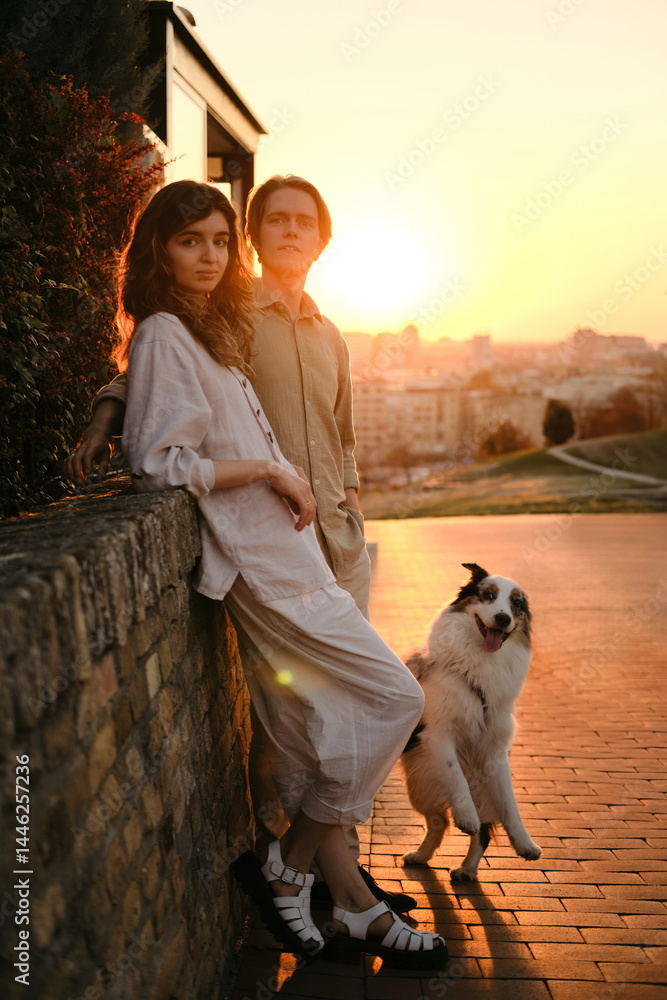 Obraz premium A young couple and their Australian Shepherd pose in the golden sunset light with the city of Novi Sad behind them. Travel in Serbia with pet
