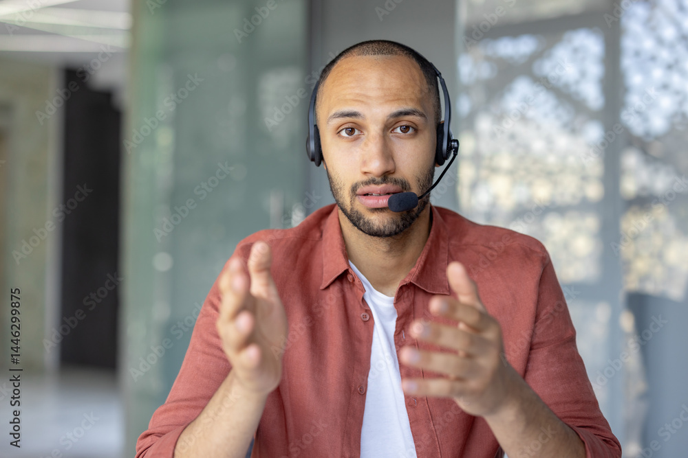 A man wearing a headset gestures while looking directly at the camera. He seems to be in a video conference call, or an online meeting.