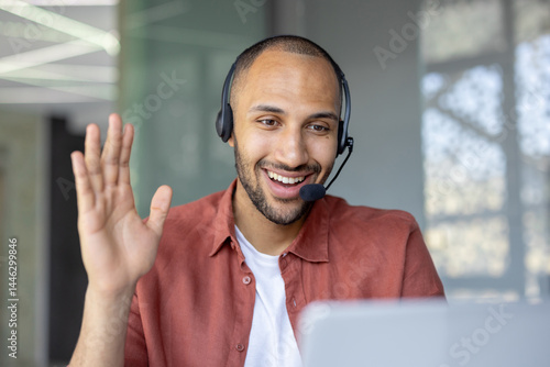 A smiling man wearing a headset waves during a video call, engaging in a friendly and professional interaction.