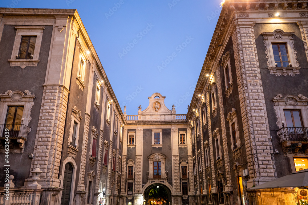 Fototapeta premium Illuminated Uzeda Gate between historic buildings in the Cathedral Square of Catania, Sicily, Italy, at dusk