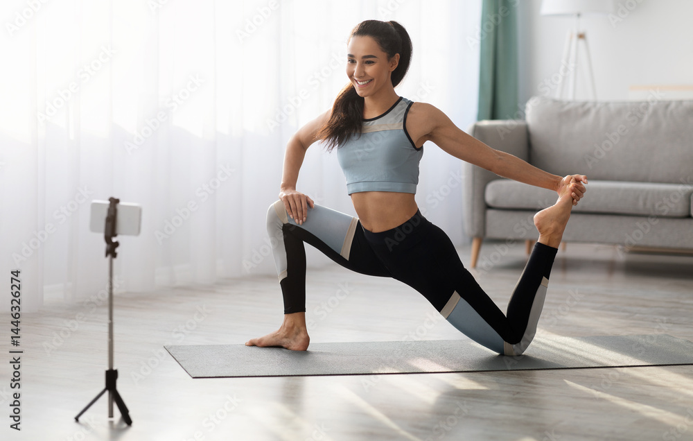 Obraz premium Positive sporty lady stretching in front of smartphone at home, copy space. Beautiful young woman fitness blogger shooting video for her followers or having personal training online