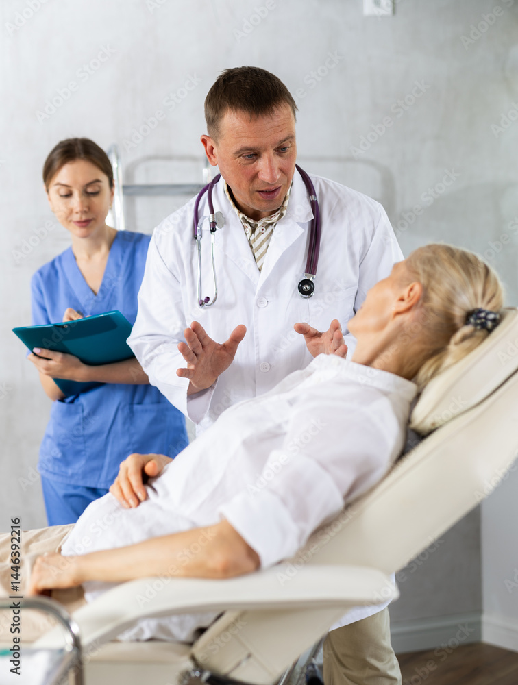 Male doctor and female assistant having conversation with elderly woman patient about need for additional diagnostic procedures, long-term and comprehensive treatment, assistant records data in report