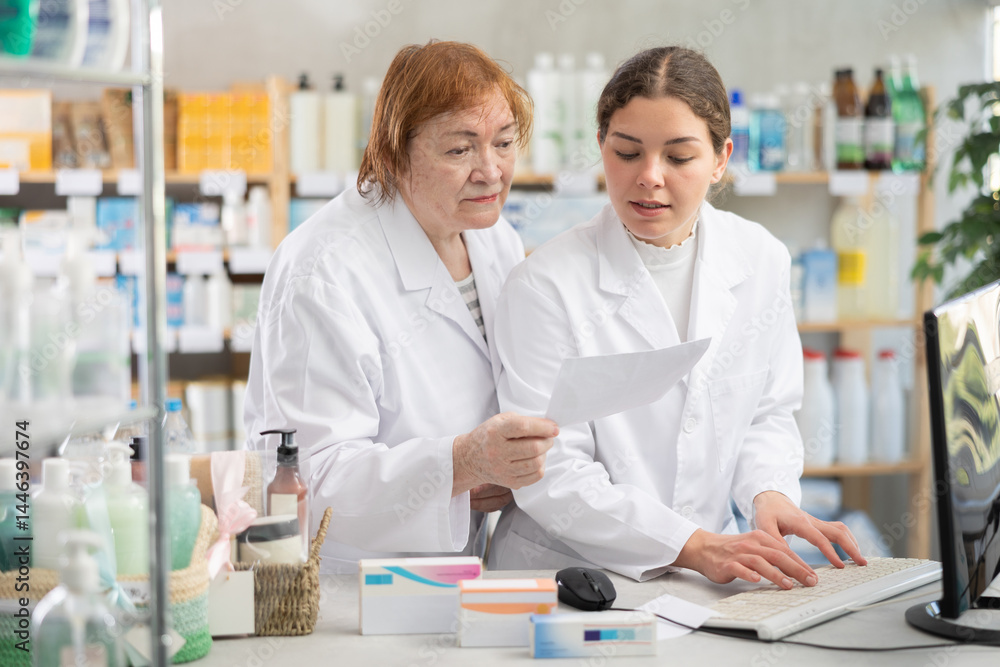 Two pharmacists stand near the counter in a pharmacy and work at a computer. Mature female pharmacist helping intern in pharmacy. Pharmacists check for medications Two pharmacists stand near the counter in a pharmacy and work at a computer. Mature female pharmacist helping intern in pharmacy. Pharmacists check for medications