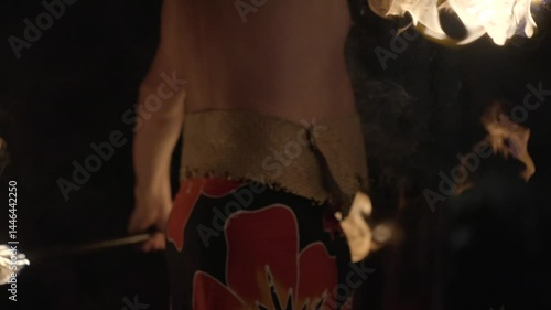 A striking close-up of a Polynesian fire dancer standing confidently with flaming torches held at his sides. Set against a deep night sky, the firelight illuminates the performer’s form.