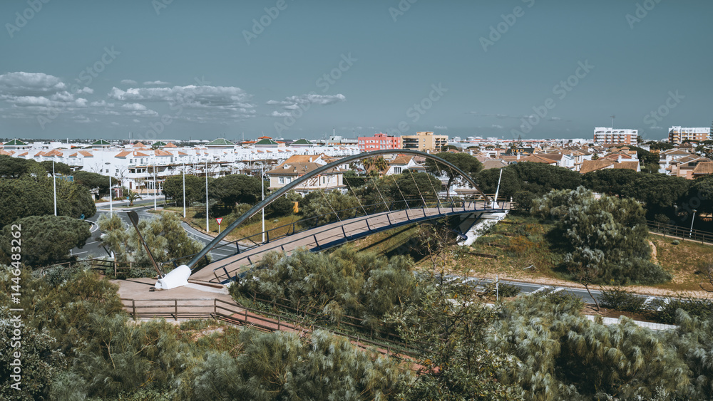 Fototapeta premium Modern pedestrian bridge with sleek metal arch crossing over green urban park, surrounded by white Mediterranean-style houses and pine trees in Punta Umbria, Andalusia, Spain