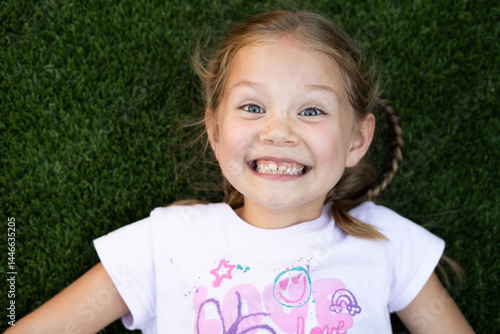 Smiling little girl with big yellow bow in her hair lying on green artificial grass, wearing black leotard, happy child portrait with natural light, outdoor lifestyle kid photo