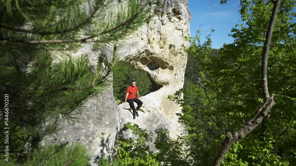 A man in a bright red shirt sits peacefully on a striking rock formation with a natural circular hole, framed by dense green forest and clear blue sky. A man in a bright red shirt sits peacefully on a striking rock formation with a natural circular hole, framed by dense green forest and clear blue sky.