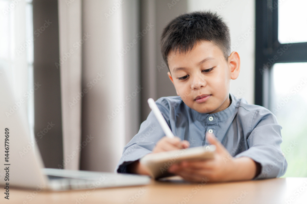 Asian young boy is focused on writing in notebook while sitting at desk with laptop nearby. scene captures moment of concentration and learning in bright, modern environment Asian young boy is focused on writing in notebook while sitting at desk with laptop nearby. scene captures moment of concentration and learning in bright, modern environment