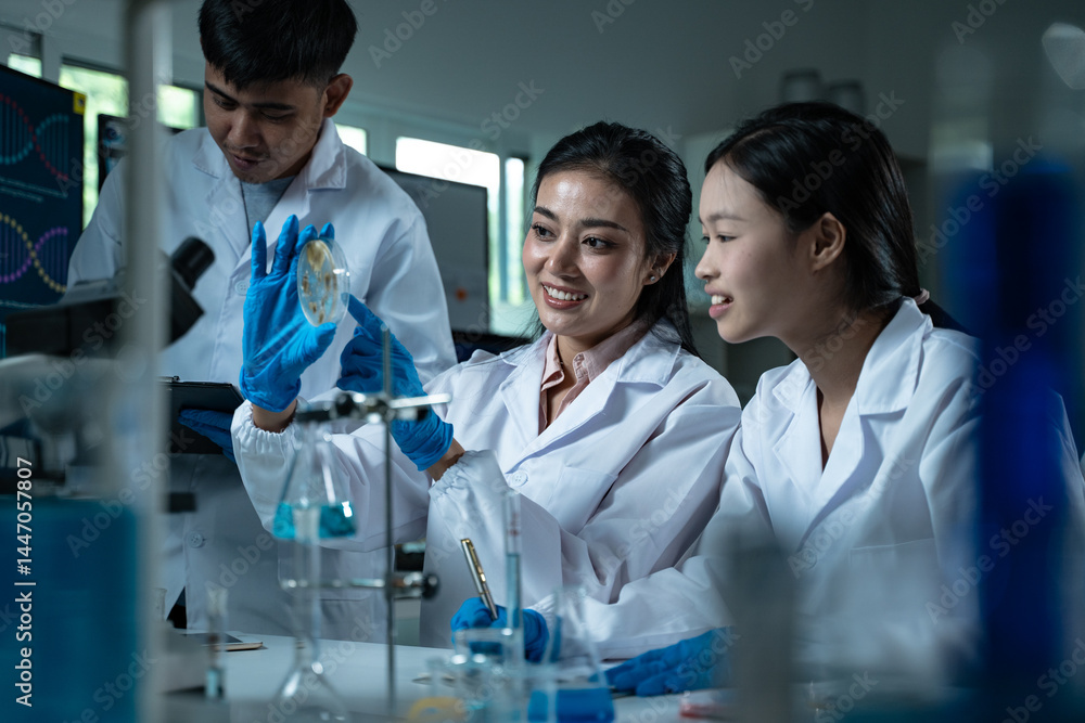 Three young scientists in lab coats working together in a laboratory examining a petri dish for microbiology biology and medical research with focus on education and innovation Three young scientists in lab coats working together in a laboratory examining a petri dish for microbiology biology and medical research with focus on education and innovation