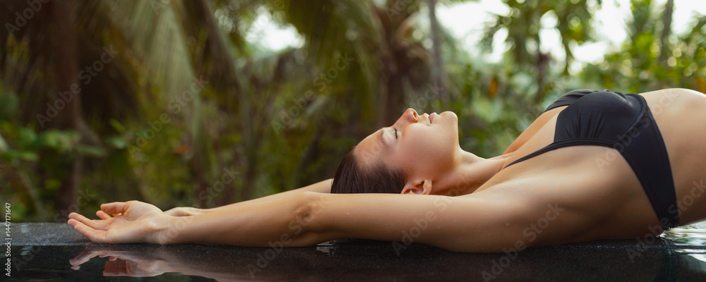 A woman in a black bikini stretches out by the edge of an infinity pool, surrounded by lush tropical greenery. Perfect for luxury travel, summer vacations, and relaxation themes, showcasing
