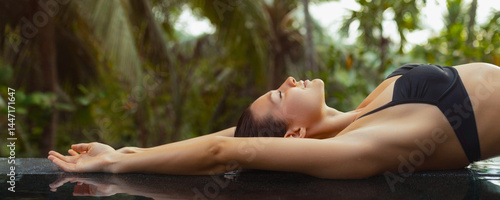 Obraz na płótnie A woman in a black bikini stretches out by the edge of an infinity pool, surrounded by lush tropical greenery. Perfect for luxury travel, summer vacations, and relaxation themes, showcasing w salonie