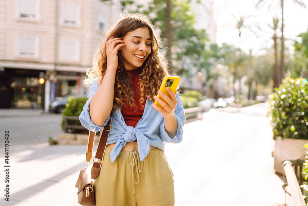 Fototapeta premium Beautiful woman with phone walking on sunny street. Tourist with phone chatting and getting new impressions outdoors. Tourism, technology concept.
