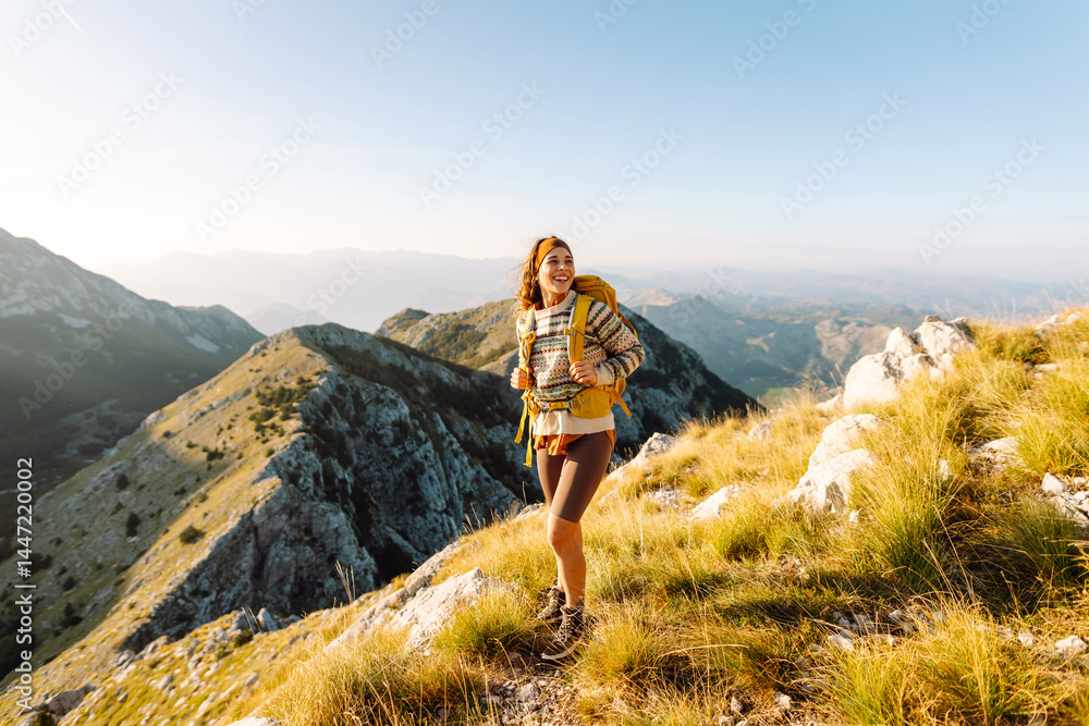 Naklejka premium Joyful woman with yellow hiking backpack in the mountains at sunset. Young female hiker enjoying the adventure and feeling freedom with mountain landscape. Wanderlust!