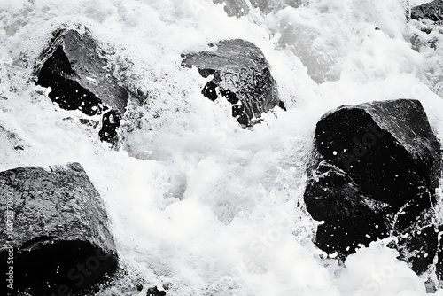 A high contrast black and white image of a rugged beach with large rocks and foamy waves crashing against them