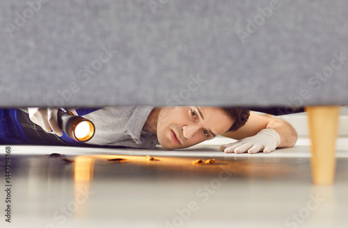 Young man worker from pest control service holding flashlight and looking under sofa at bugs or cockroaches crawling on floor. Roaches at home, house insects concept