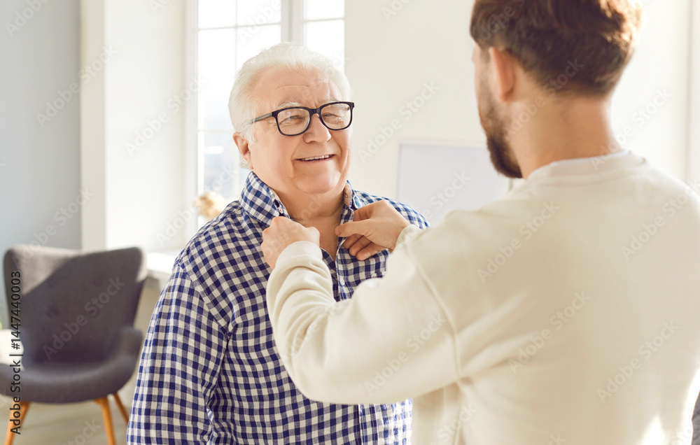 Obraz premium Adult son helping his senior father to put on his clothes. Young man buttons up the shirt of his happy, cheerful, smiling old dad. Family, older age, care concept