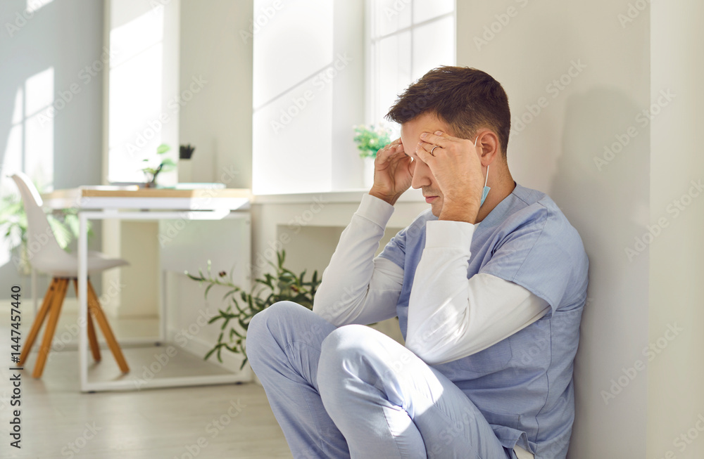 Stressed man nurse or doctor in blue scrubs feeling tired, exhausted and completely burnt out after long difficult work shift under constant pressure sitting by wall on floor holding hands on forehead