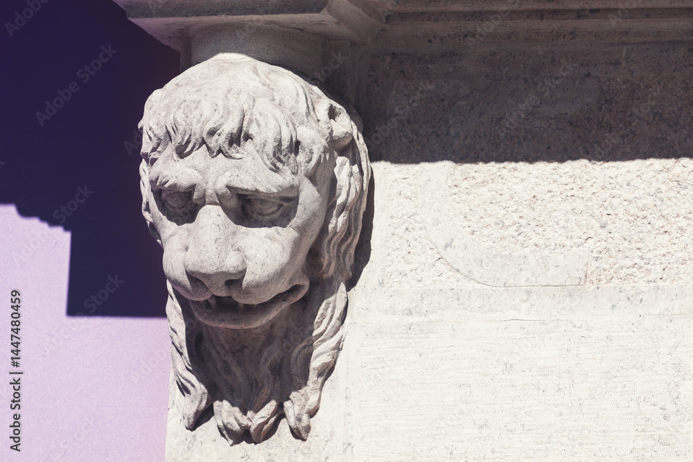 Stone carving of lion head, with detailed features and fierce expression, adorns corner of a building's column. Sculpture casts a shadow on colored stone, with a purple wall visible in background