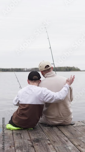 Happy son hugs his dad while fishing on a wooden pier. Father teaches his child to fish. Concept of family and fatherhood. Vertical video.