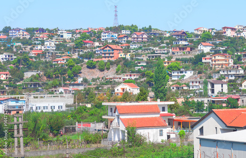 Suburb in Albania covers a hilly area with houses, mostly with tiled roofs, under a clear blue sky. White house with a red roof sits in foreground, while a telecommunications tower looms in distance