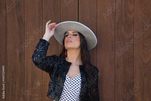 Portrait of young woman, beautiful, brunette, flamenco dancer, wearing beautiful dress with jacket and hat, posing with wooden door in background. Flamenco concept, dancer, typical, Spanish, Seville.