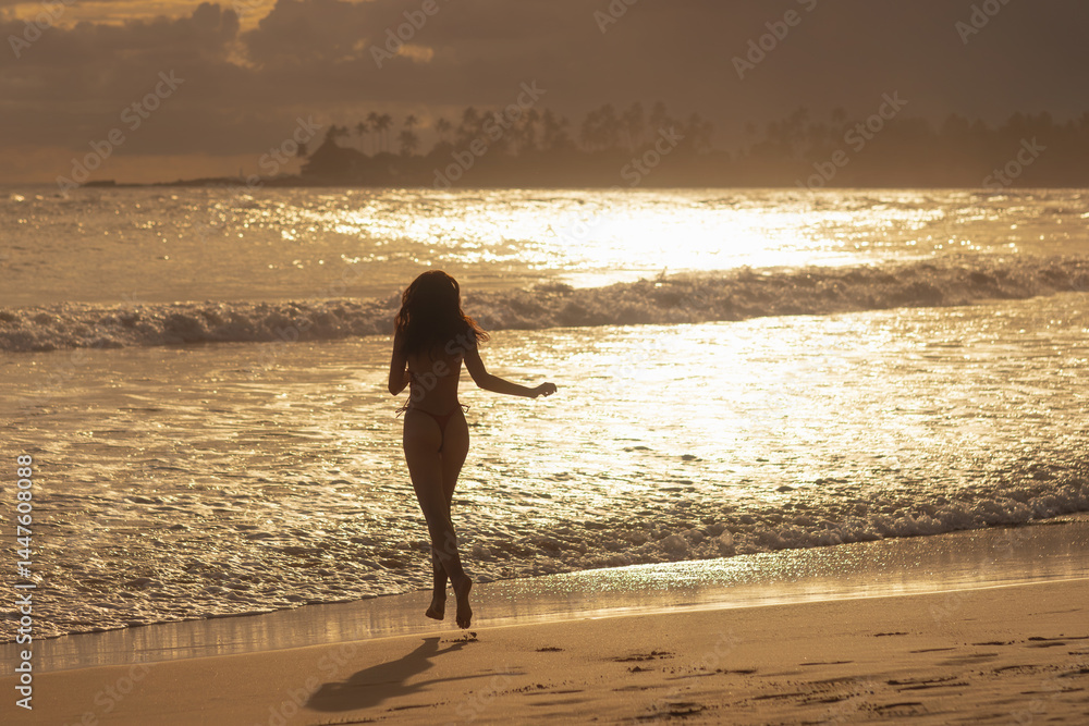 Backlit silhouette of a woman running joyfully on the beach as golden sunset reflects on the ocean - concept for freedom, tropical travel, sunset beauty, wellness, and inspirational lifestyle visuals.