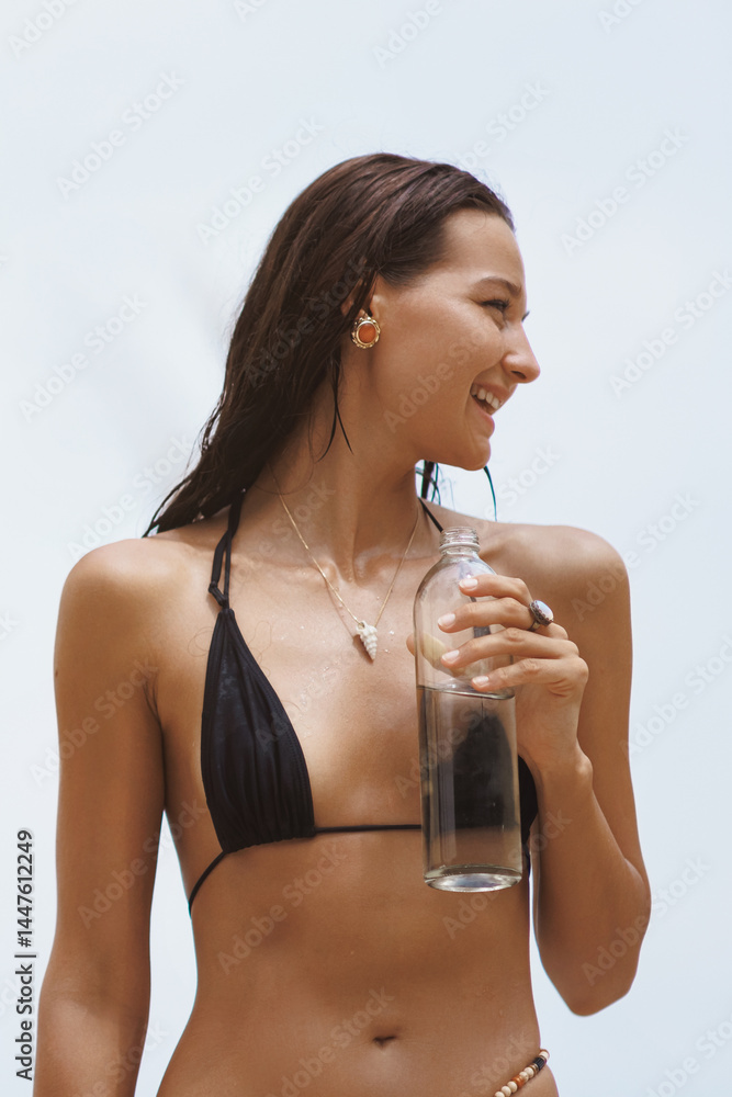 Close-up of a cheerful Asian woman in a black bikini holding a glass bottle of water, smiling brightly against a tropical sky. Ideal for skincare, hydration, wellness, and summer promotions.