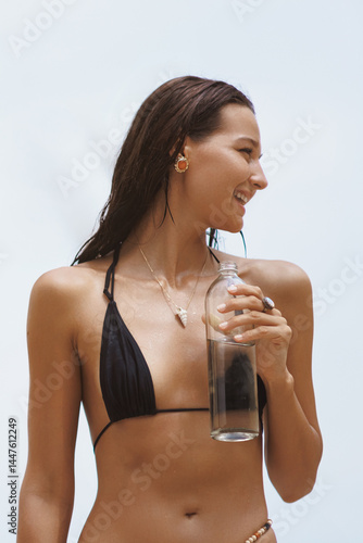 Obraz na płótnie Close-up of a cheerful Asian woman in a black bikini holding a glass bottle of water, smiling brightly against a tropical sky. Ideal for skincare, hydration, wellness, and summer promotions. w salonie