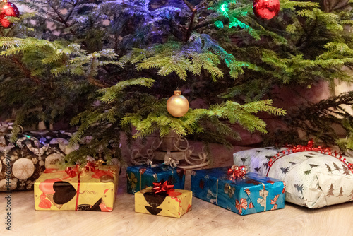 
The bottom of a Christmas tree with presents wrapped in colorful paper and decorated with bows, placed on the floor. The tree is decorated with red and gold balls.