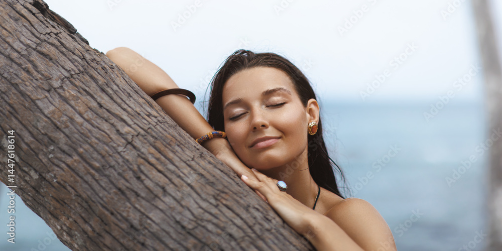 Close-up of a peaceful Asian woman resting her face on a palm tree with eyes closed by the ocean. Ideal for wellness, relaxation, skincare and beauty-related promotional concepts.