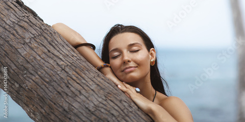 Obraz na płótnie Close-up of a peaceful Asian woman resting her face on a palm tree with eyes closed by the ocean. Ideal for wellness, relaxation, skincare and beauty-related promotional concepts. w salonie