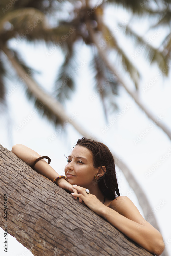 Tanned Asian woman resting against a palm tree and looking thoughtfully away with a serene expression. Perfect for skincare, tropical travel, beauty and peaceful lifestyle promotions.
