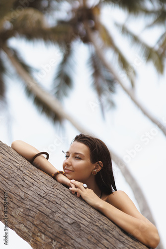 Obraz na płótnie Tanned Asian woman resting against a palm tree and looking thoughtfully away with a serene expression. Perfect for skincare, tropical travel, beauty and peaceful lifestyle promotions. w salonie