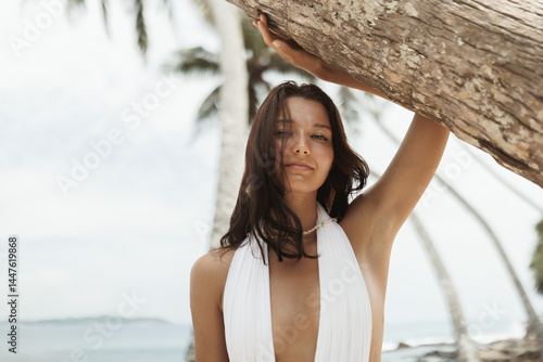 Obraz na płótnie A woman in a plunging white swimsuit leans against a palm tree on a tropical beach. She gazes into the distance with a serene expression. The ocean and additional palm trees create a beautiful and w salonie