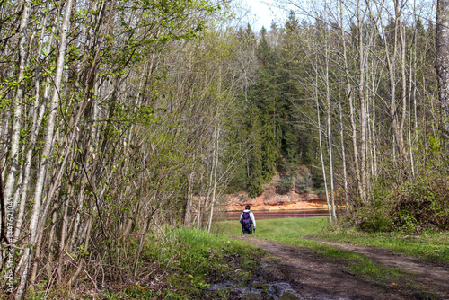 
A man in dark clothing with a backpack walking along a dirt road in a wooded area, with trees with newly emerging leaves in the foreground and dark conifers and a reddish rock in the background.
