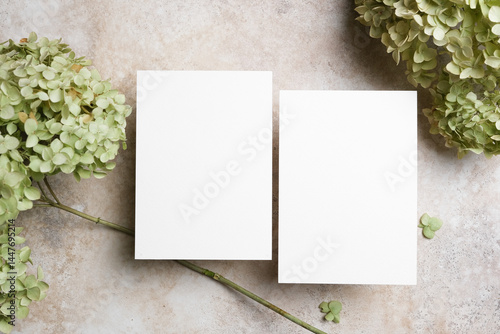 Double blank wedding invitation cards mockup on a neutral stone surface surrounded by pale green hydrangea flowers, ideal for elegant greeting or flyer card designs