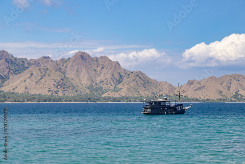 Traditional wooden boat sailing on clear turquoise waters with arid hills and mountain range in the background under a bright sky. Scenic view from East Nusa Tenggara, Indonesia.