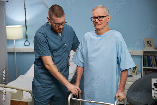 Senior man being assisted by healthcare professional while standing with walker in hospital room. Calm demeanor of both individuals in clinical setting