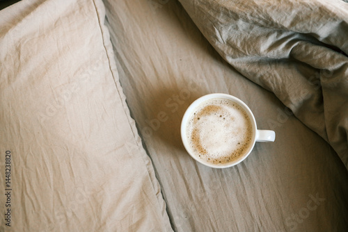 Fotografie White ceramic cup of frothy cappuccino resting on soft beige bedding