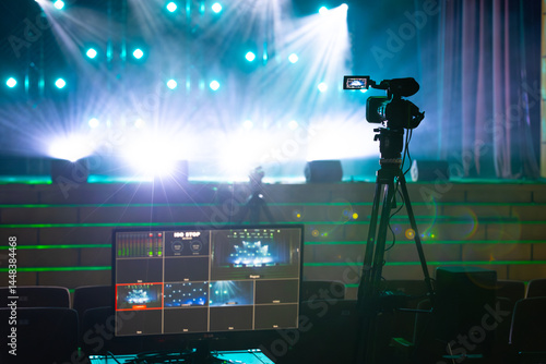 A professional video camera and live monitor in front of a brightly lit concert stage with blue and green lighting effects.