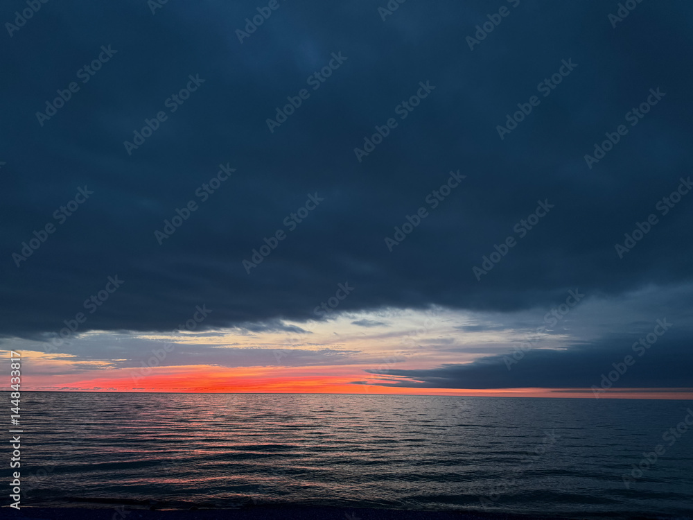 Obraz premium Cloudy sky over a calm sea at sunset with red and orange colors on the horizon. The majority of the sky is covered in dark clouds.