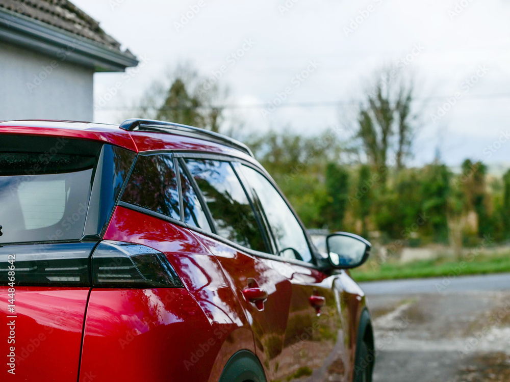 Red car parked outside house, small rural road and country side green area in the background. Living out of town or a village concept. Transportation and commute between work and home theme.