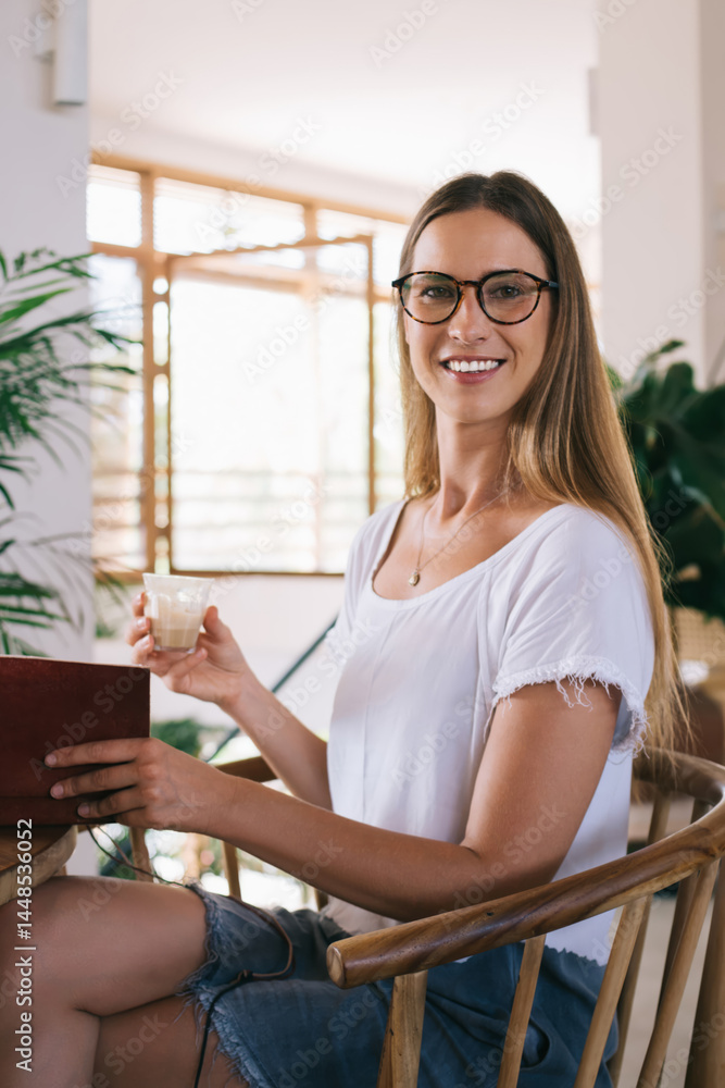 Positive woman drinking beverage while working with diary and laptop