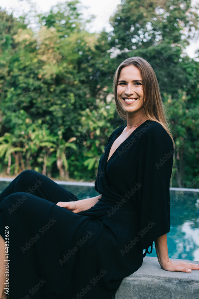 Happy young woman sitting on pool margin in tropical garden